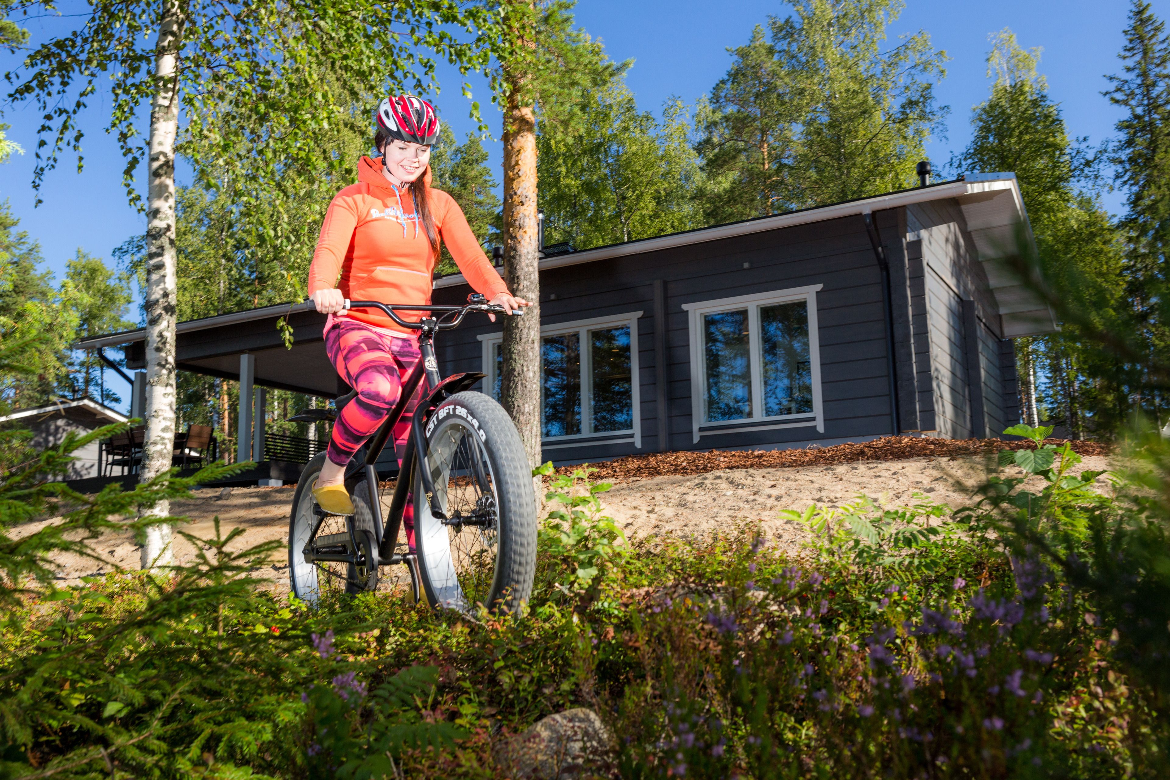 A person riding a bicycle in front of a modern house with a covered porch and a garden.