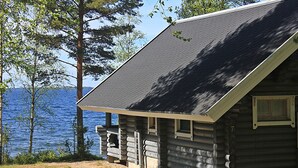 A log cabin with a sloped roof, a window, and a door, situated near a body of water with trees in the background. - Karelia Country Cottages (Kitee)