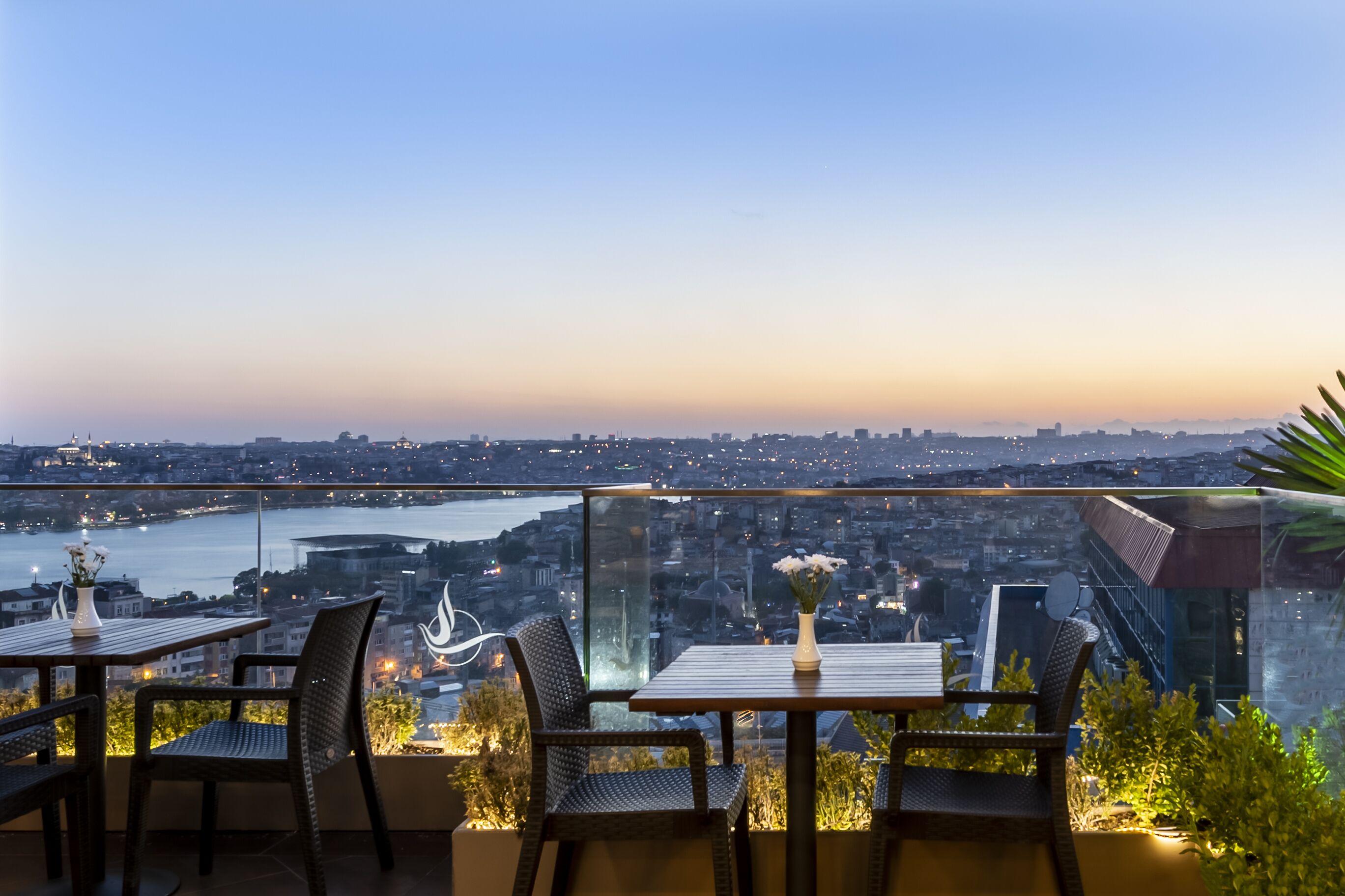 Une terrasse avec une table et des chaises, d’où l’on peut admirer un panorama sur la ville et un plan d’eau au crépuscule.