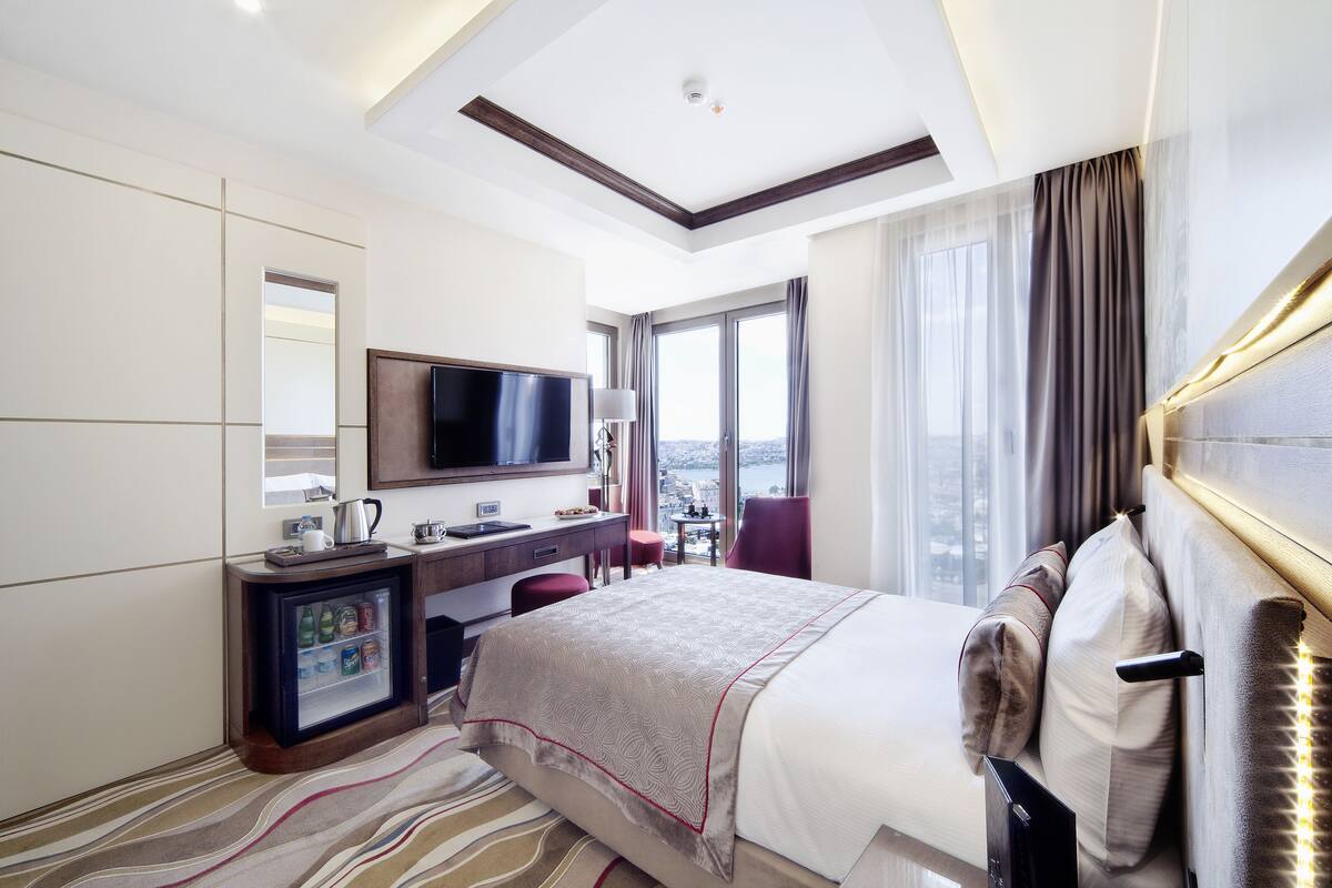 A hotel room with a bed, a desk, a television, a minibar, and a city view.