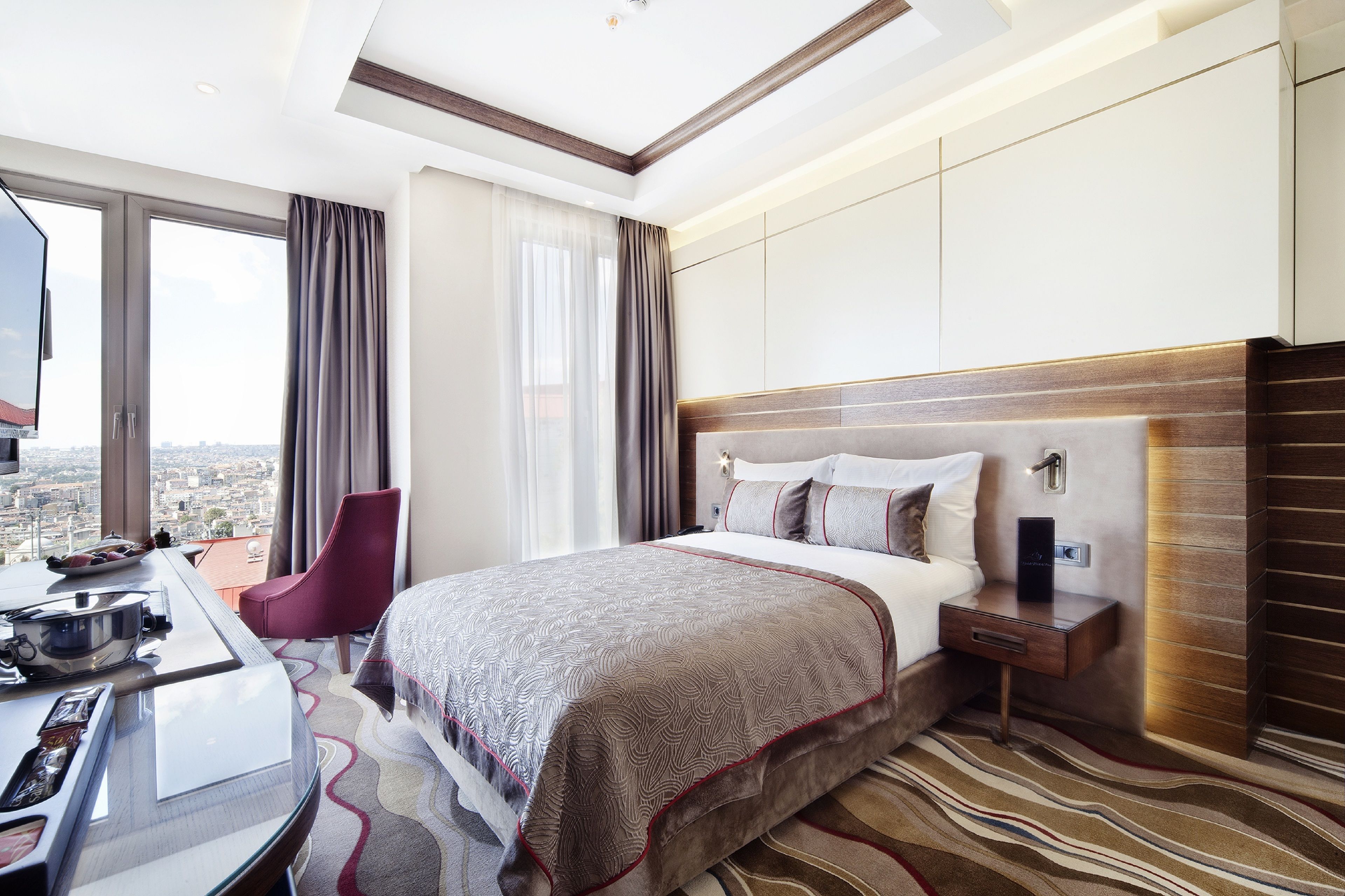 Hotelkamer met een groot bed, een bureau met een laptop, een stoel en uitzicht op de stad door grote ramen.