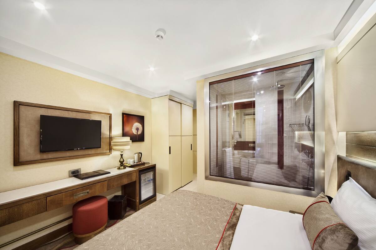 A hotel room with a large bed, a flat-screen TV mounted on the wall, a desk with a lamp, and a large window with blinds.