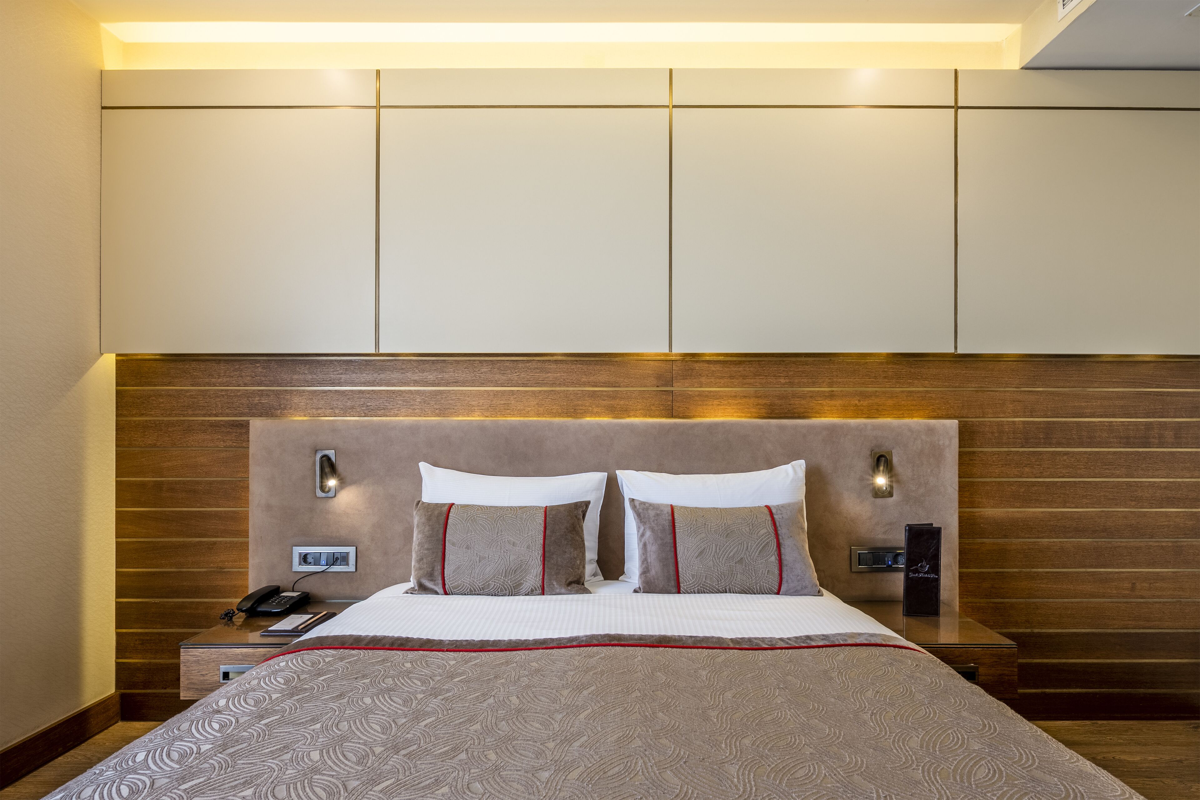 A hotel room with a large bed, wooden headboard, bedside lamps, a nightstand with a phone and a remote control, and a wall-mounted panel.