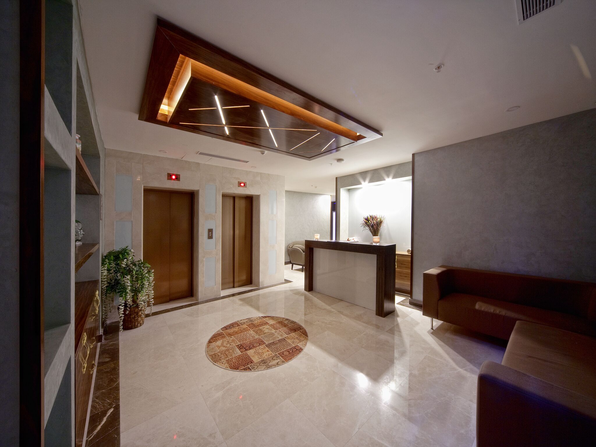 A modern lobby with a reception desk, two elevators, a patterned rug, and a seating area.