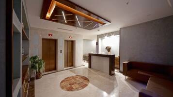 A modern lobby with a reception desk, two elevators, a patterned rug, and a seating area.