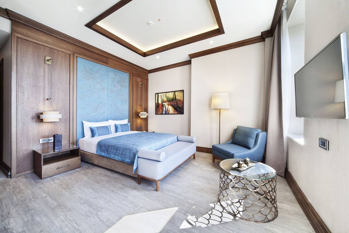 A modern hotel room with a large bed, a seating area with a chair, a TV, and a painting on the wall.