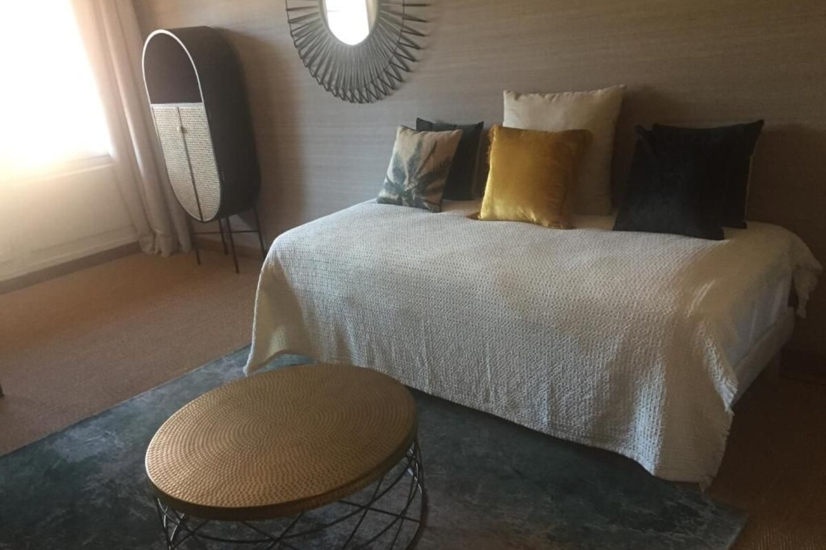 A bedroom with a bed, a round woven stool, a tall cabinet, and a wall-mounted mirror.