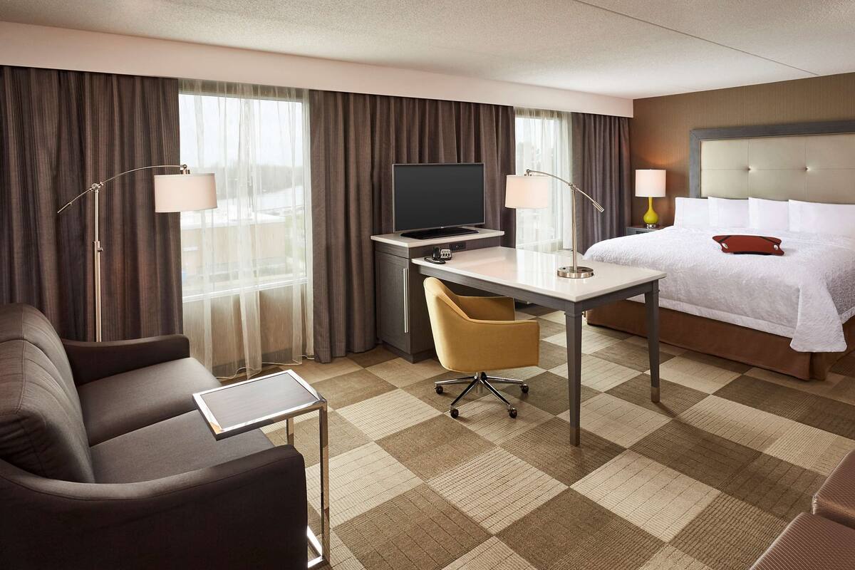 A hotel room with a bed, desk, two chairs, a TV, and lamps.