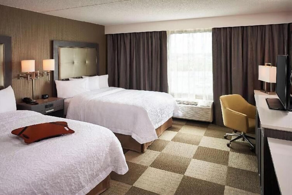 Ein Hotelzimmer mit zwei Betten, einem Schreibtisch, einem Stuhl, einem Fernseher und einem Fenster mit Vorhängen.