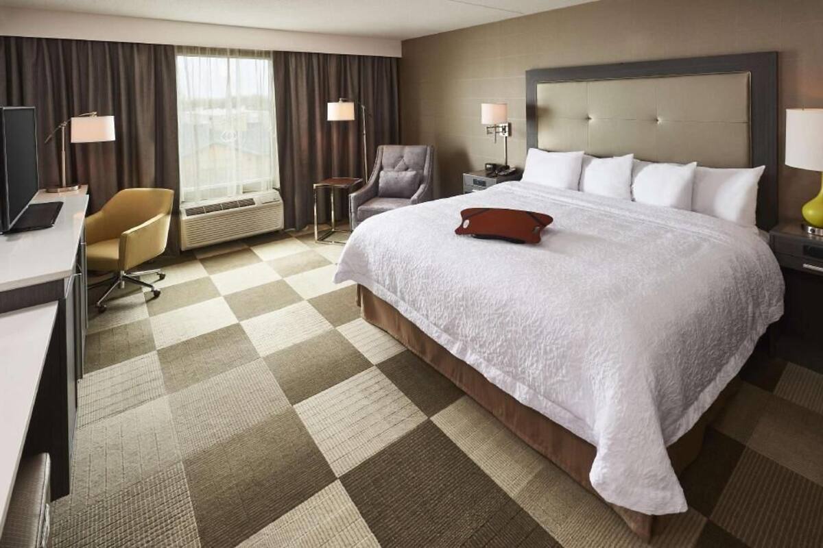 A hotel room with a large bed, a desk, a chair, a lamp, and a window.