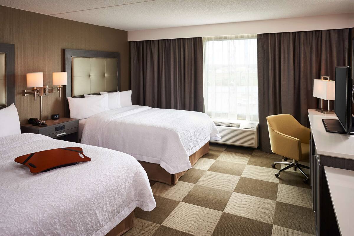 A hotel room with two beds, a desk, a chair, a TV, and a window with curtains.