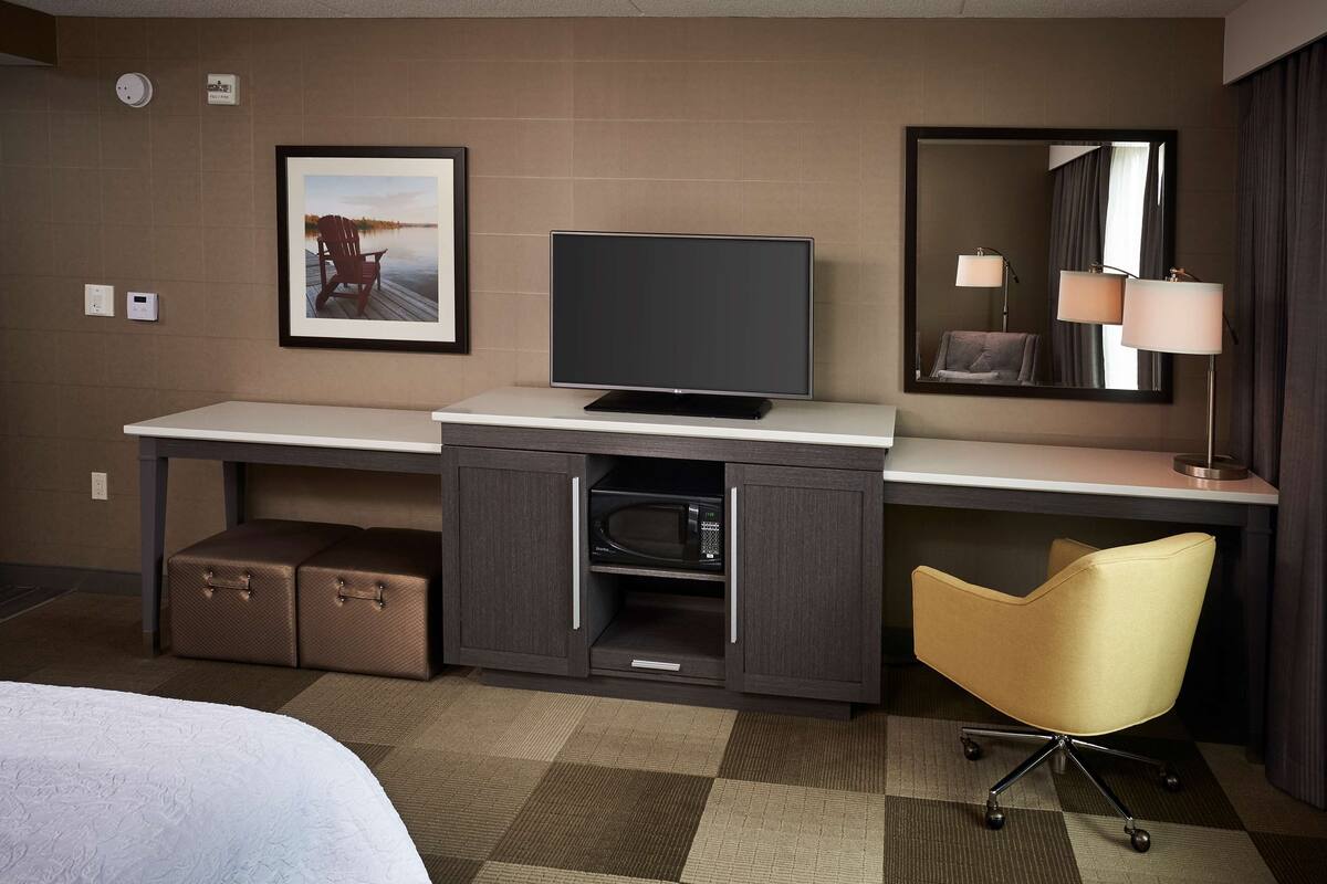 A hotel room with a bed, a desk with a television, a microwave, a chair, and a framed picture on the wall.