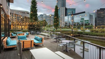 Een terras buiten met houten meubels en lichtjes, met uitzicht op een rivier en moderne gebouwen.