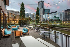 An outdoor seating area with wooden furniture and string lights, overlooking a river and modern buildings. - Courtyard by Marriott Amsterdam (Amsterdam)