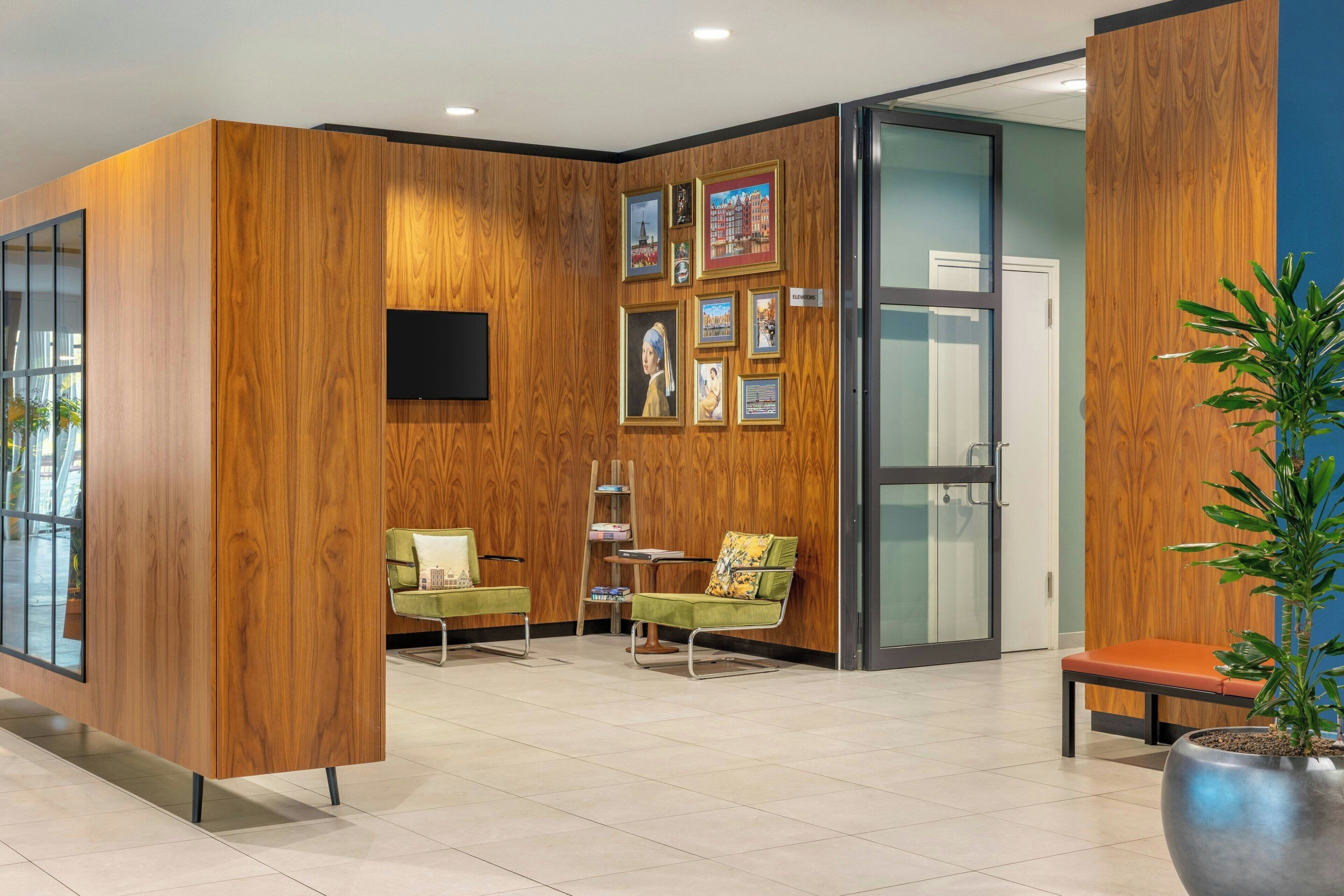 Un área de recepción con paneles de madera, un televisor de pantalla plana, cuadros enmarcados, una silla verde, un banco y una planta en maceta.