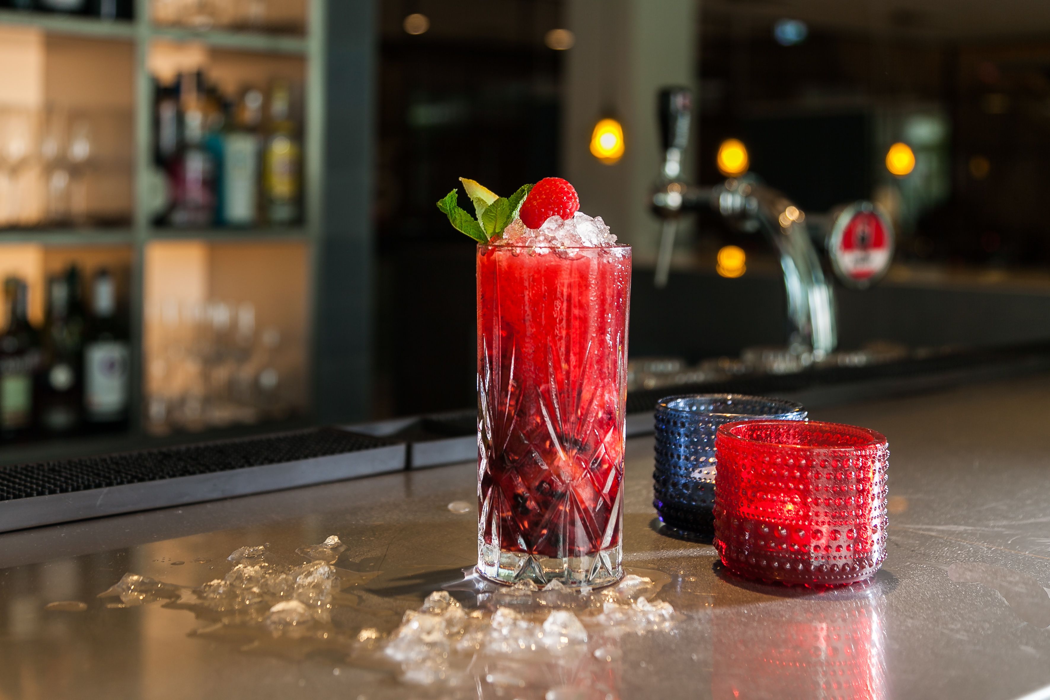 Un bar con un vaso alto de bebida roja decorado con una fresa y menta, cubitos de hielo y dos vasos rojos sobre la barra.