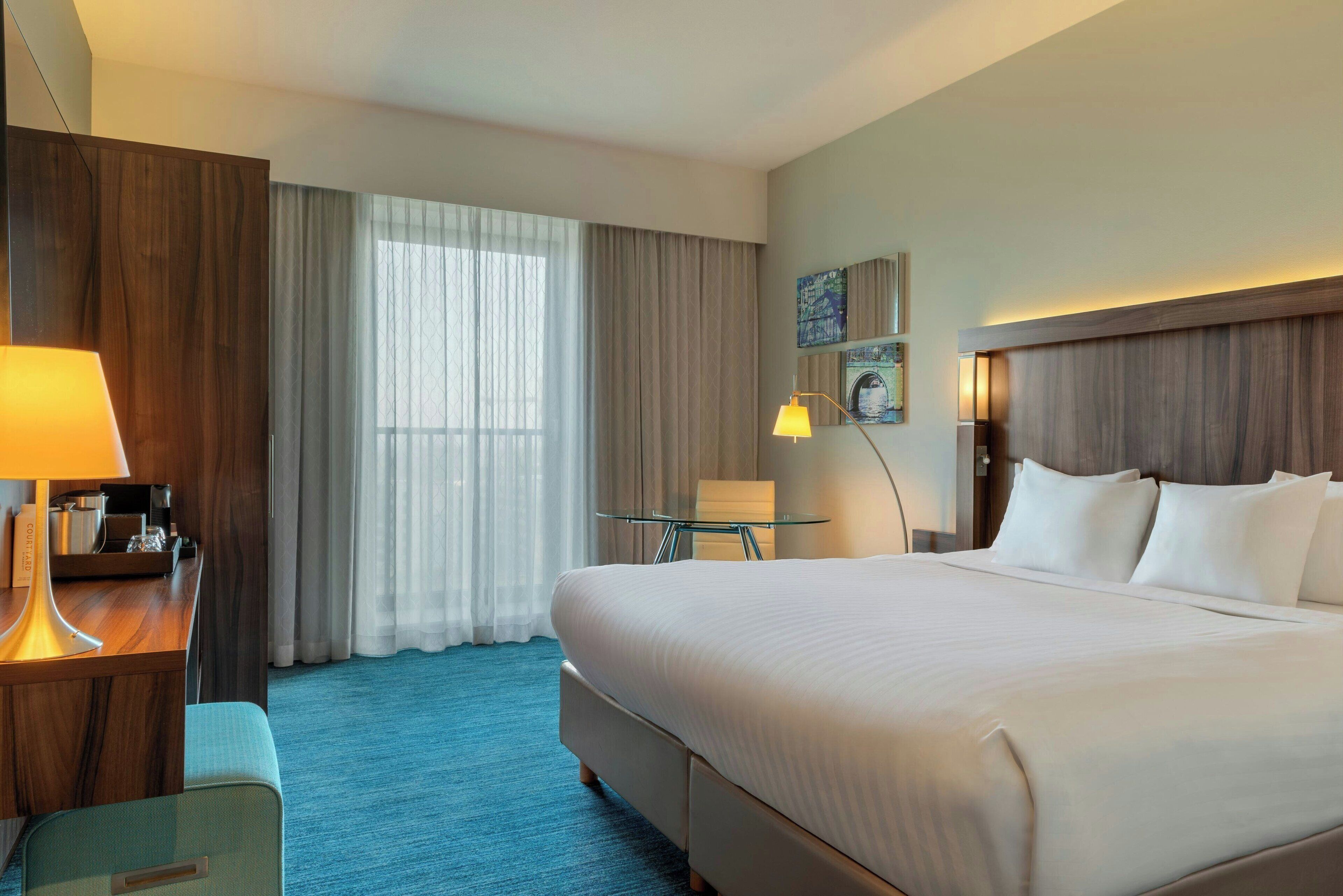 A hotel room with a large bed, a desk with a lamp, a small table, and a chair.