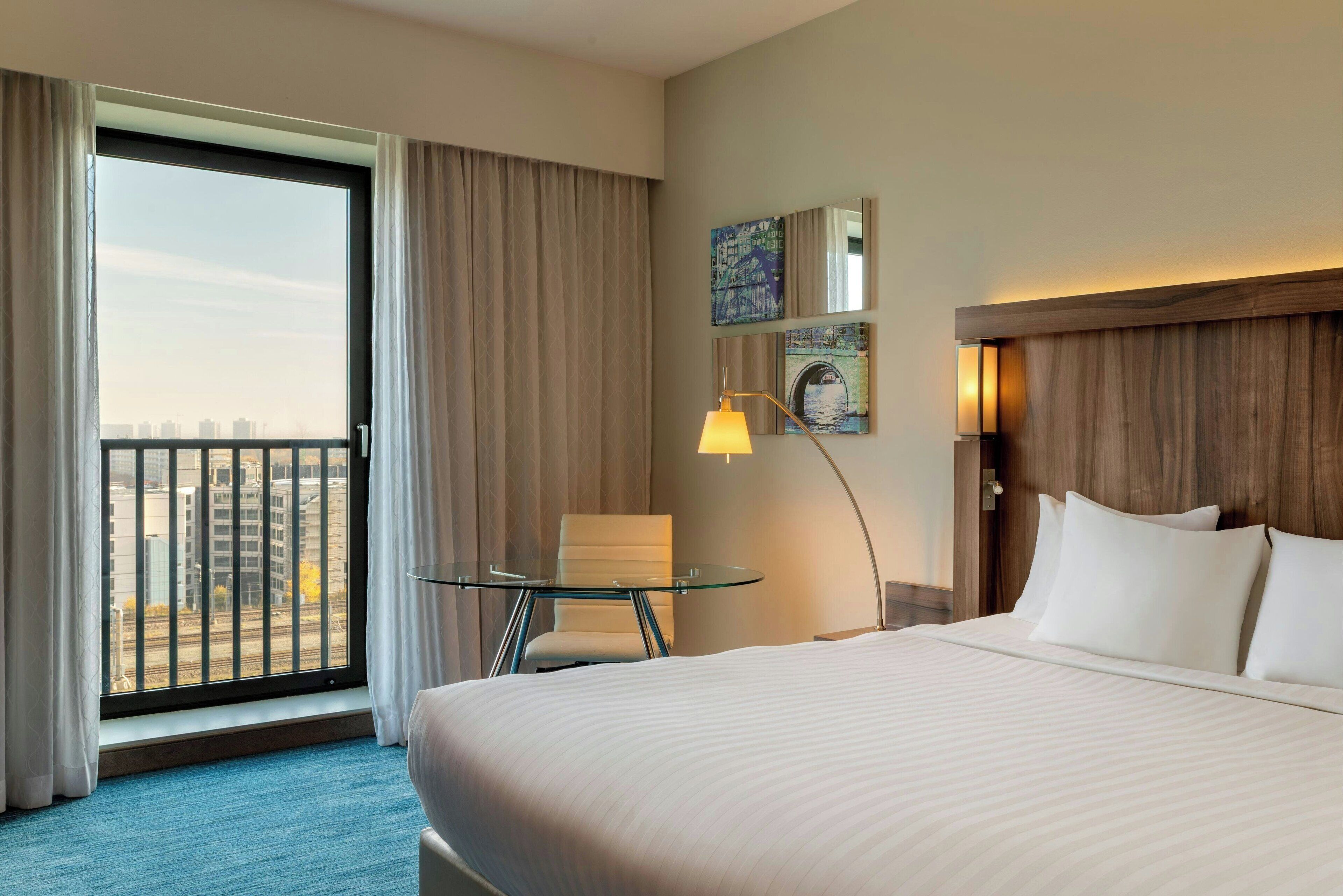A hotel room with a large bed, a desk with a chair, a view of the city through a large window, and artwork on the wall.