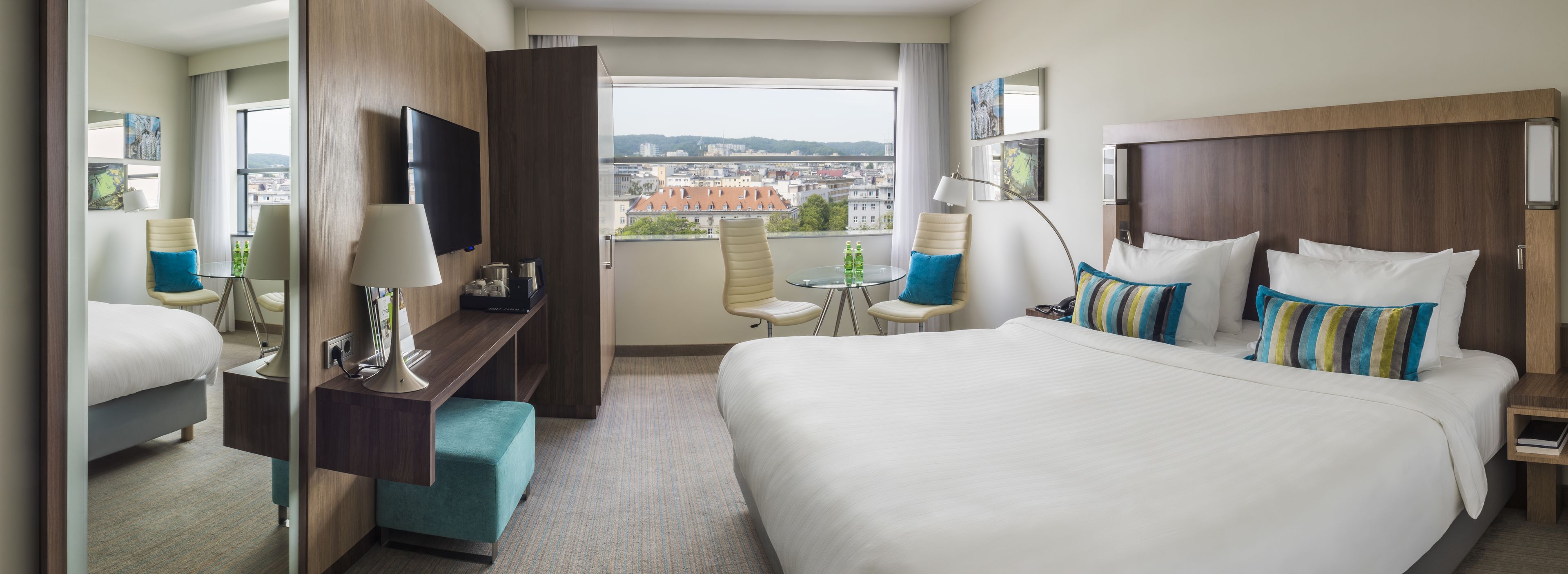 A hotel room with a large bed, a desk with a lamp, a chair, a small table with two bottles, and a TV on the wall.