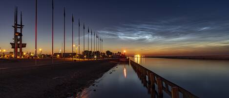 A waterfront promenade at dusk with a calm sea, a wooden pier, and street lamps.