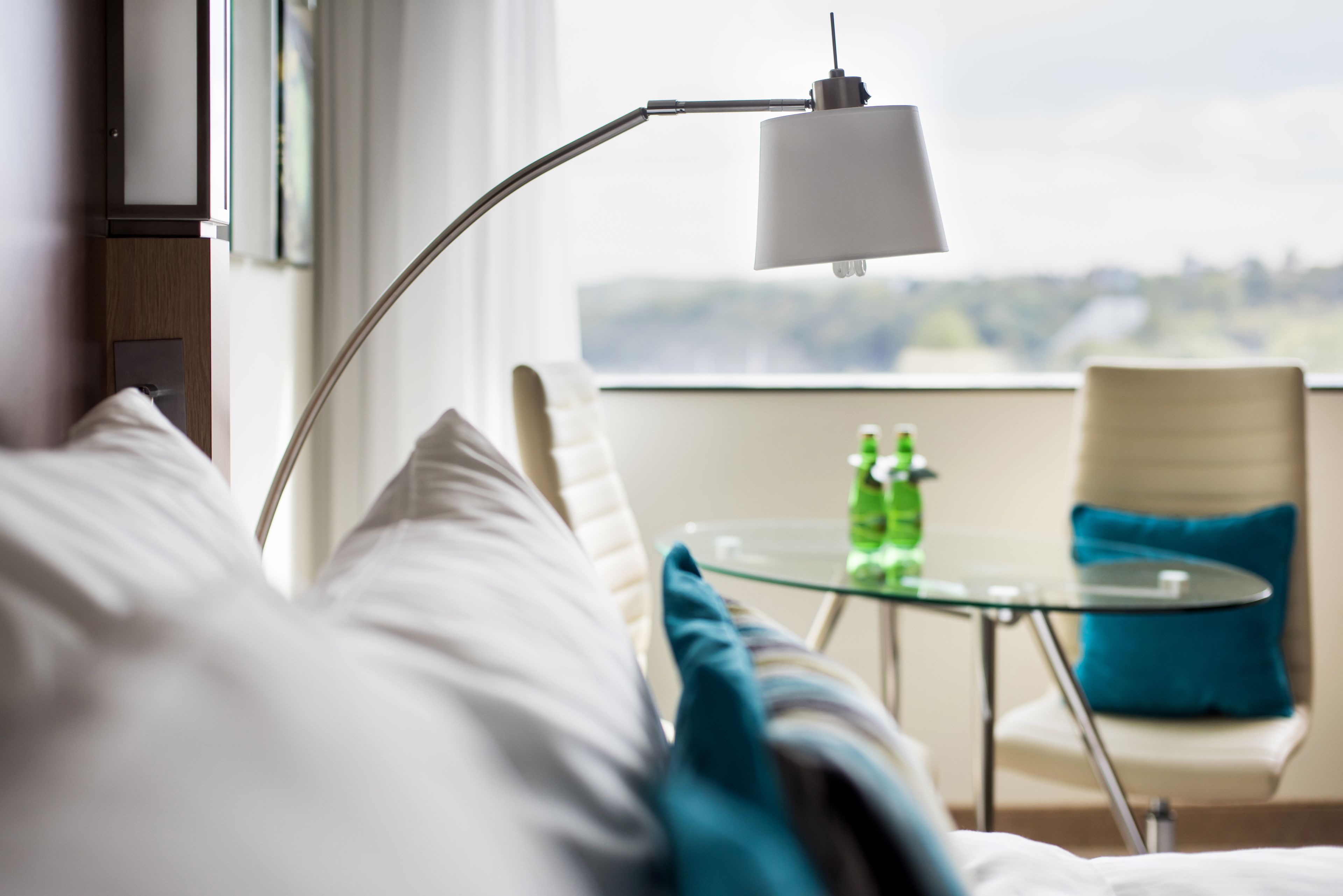 A hotel room with a bed, a glass table with two bottles, a chair with a blue pillow, and a lamp.