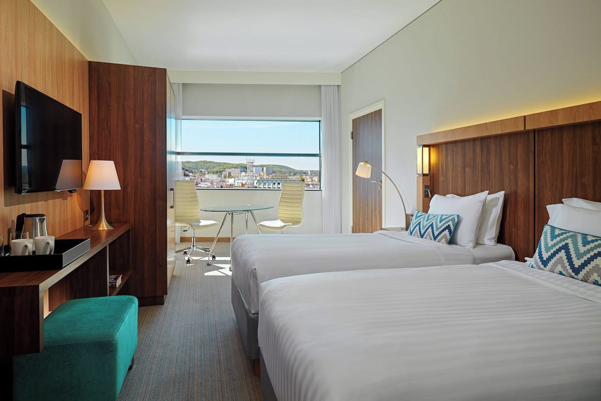 A hotel room with two beds, a TV, a desk with a lamp, and a view of the city.