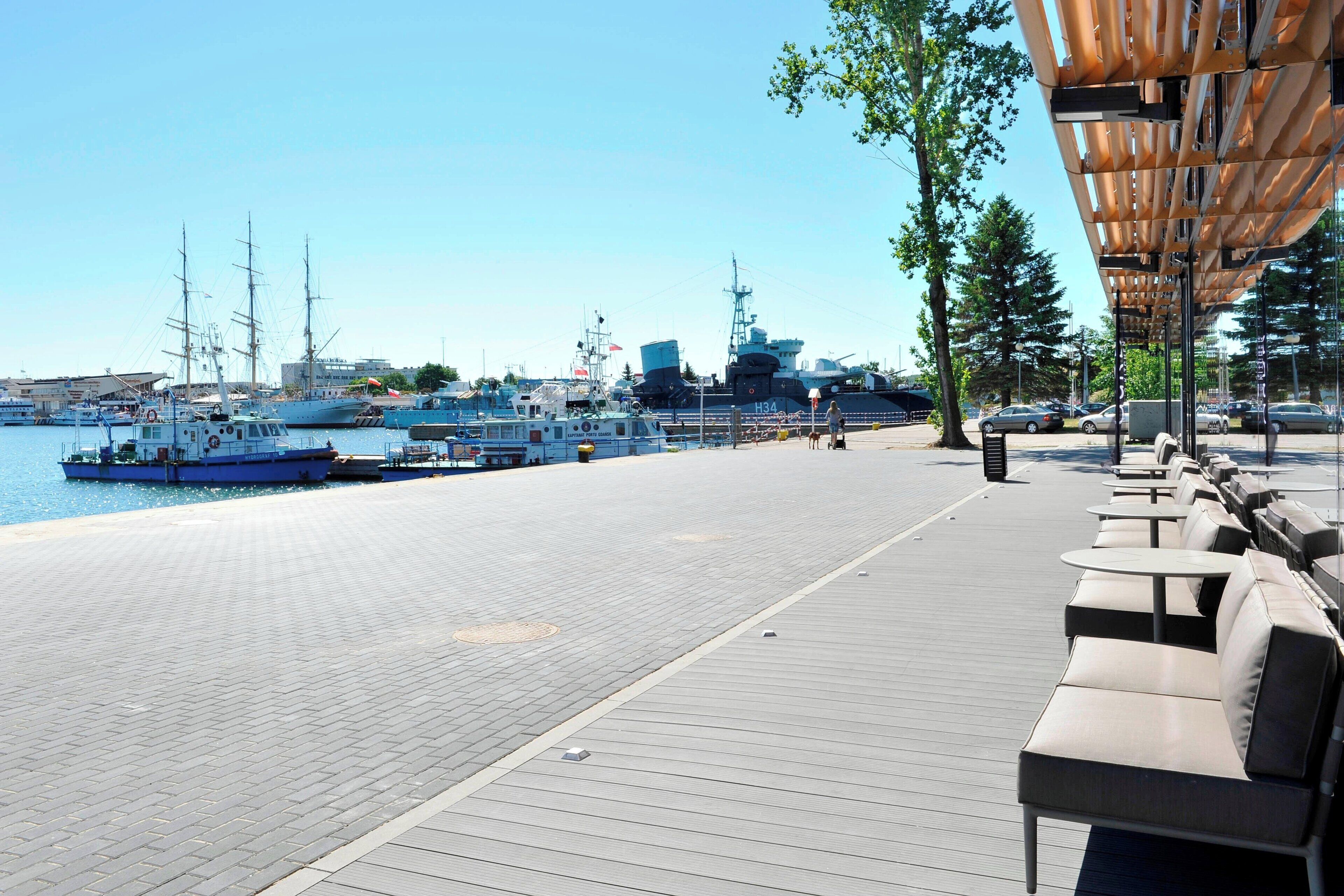Eine Uferpromenade mit Sitzgelegenheiten im Freien und Blick auf mehrere Boote, die in einer Marina vor Anker liegen.