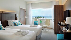 A hotel room with two beds, a desk, a chair, a small table with bottles, and a view of the sea.
