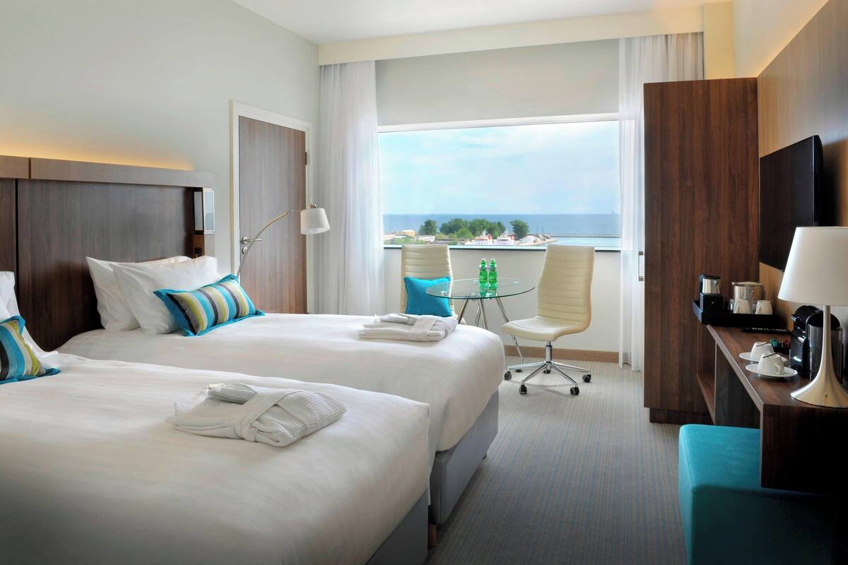 A hotel room with two beds, a desk, a chair, a small table with bottles, and a view of the sea.