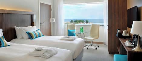 A hotel room with two beds, a desk, a chair, a small table with bottles, and a view of the sea.
