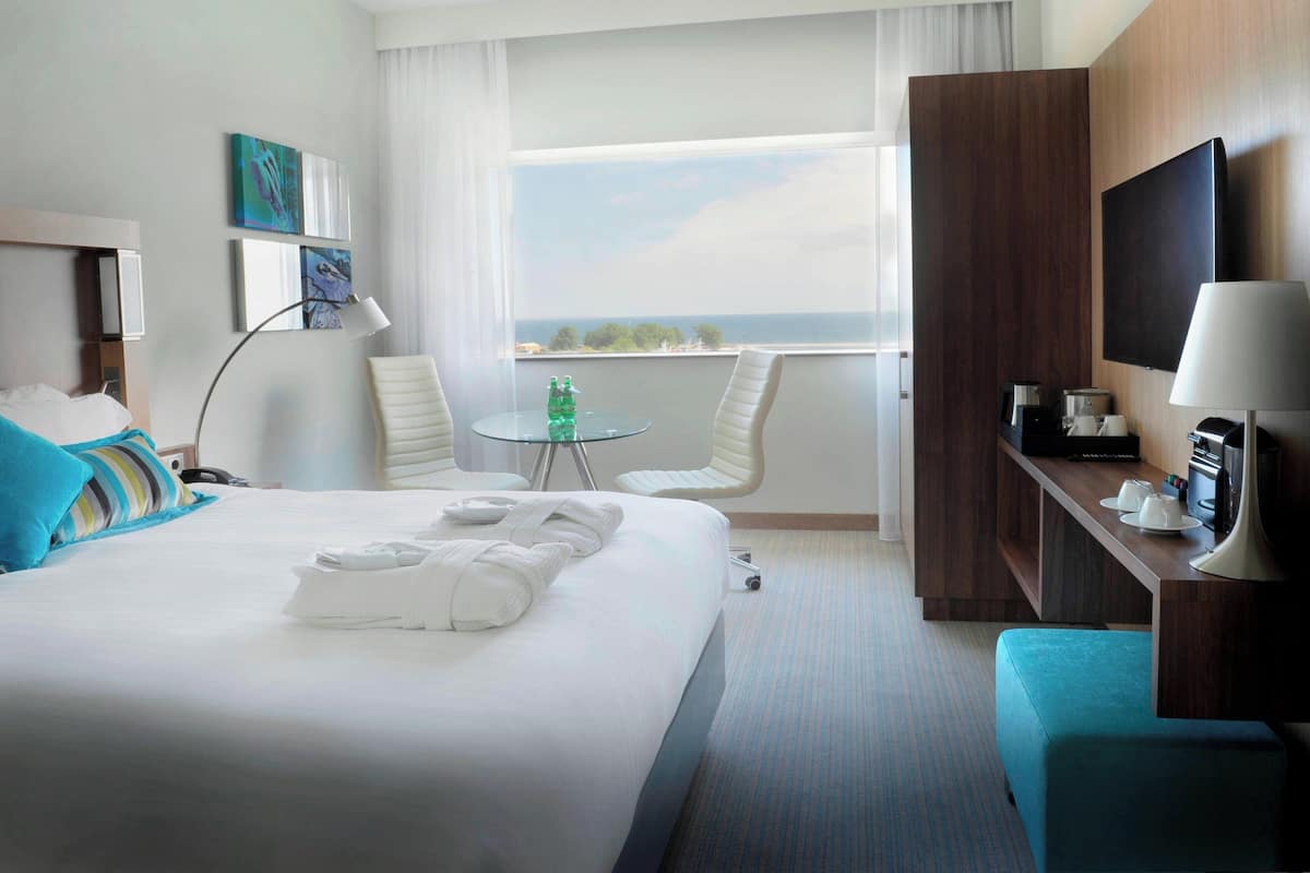 A hotel room with a large bed, a desk with a TV, a small table with chairs, and a balcony with a view.