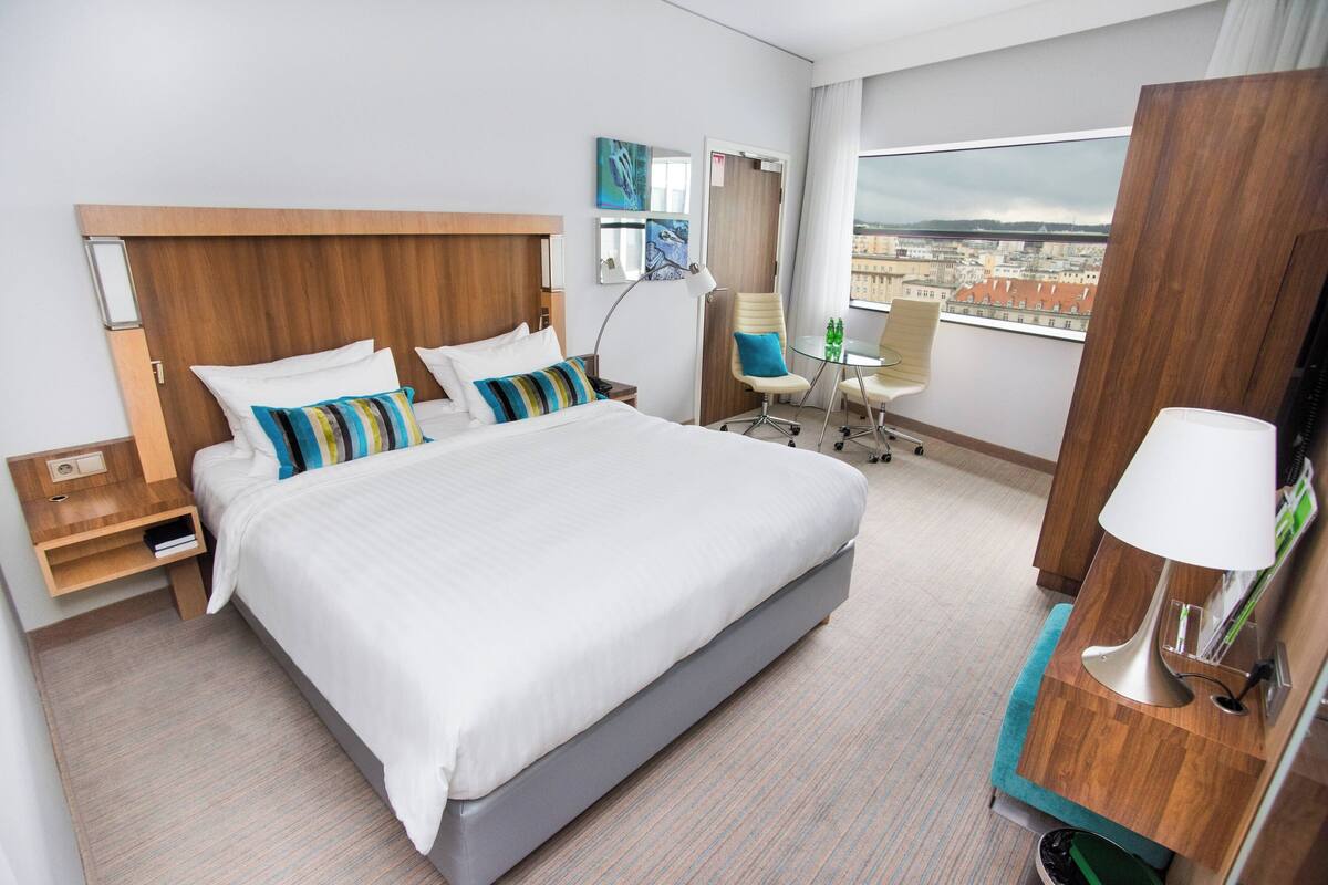 A modern hotel room with a large bed, a desk, a chair, and a view of the city.