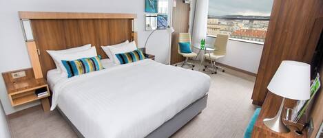 A modern hotel room with a large bed, a desk, a chair, and a view of the city.