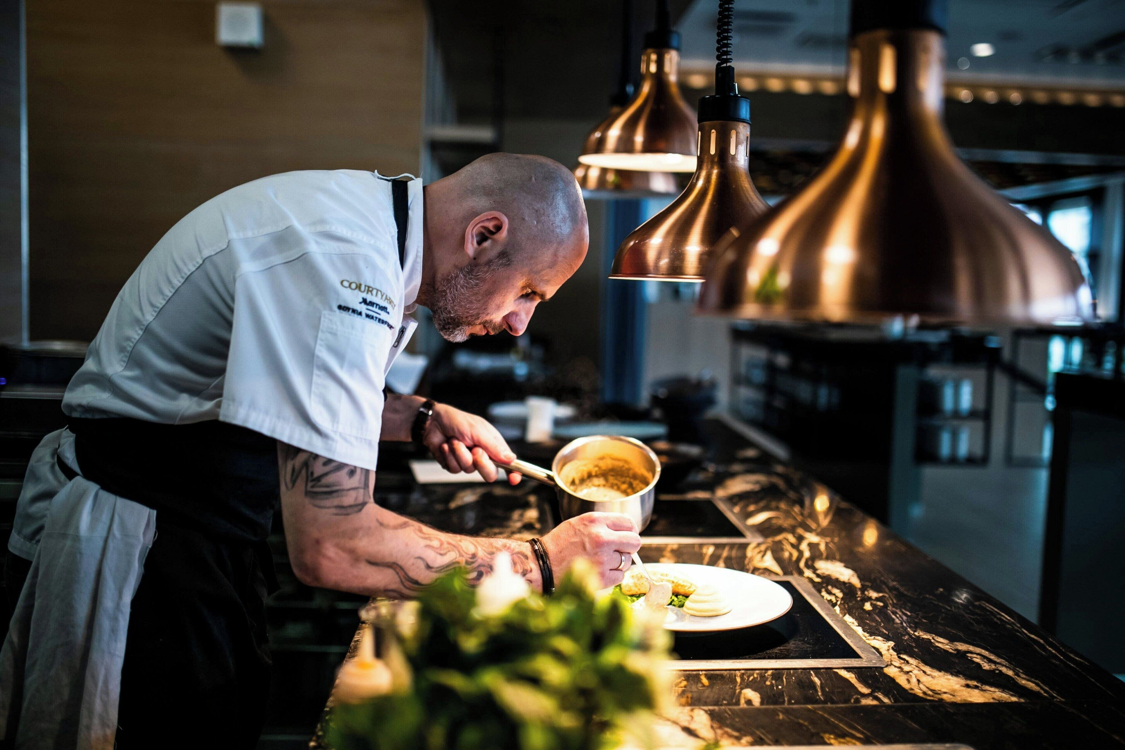 Un chef préparant des plats dans une cuisine moderne, dotée de luminaires suspendus en cuivre et d’un plan de travail en marbre.