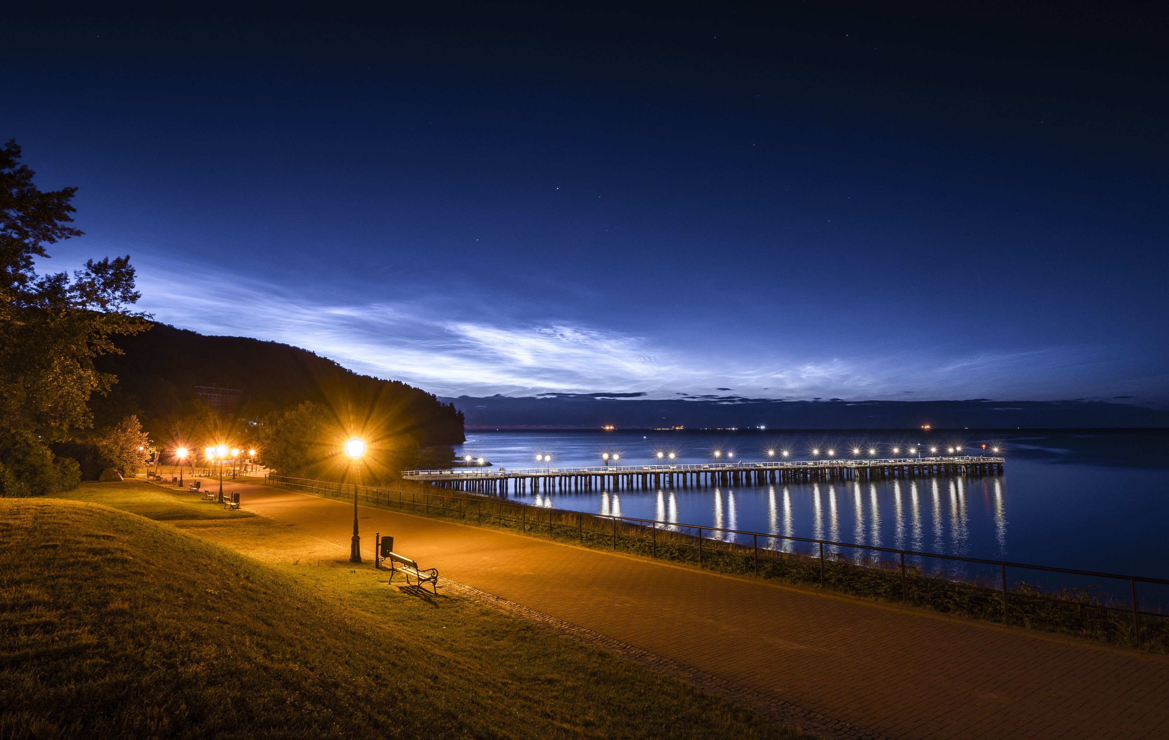 Vue nocturne d’un parc au bord de l’eau, avec une promenade dallée, des bancs et une jetée s’avançant dans des eaux calmes.