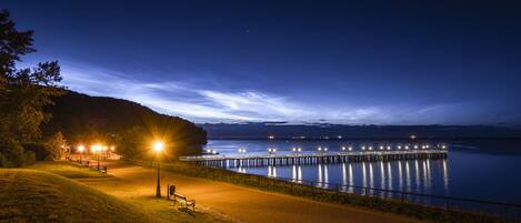 Vue nocturne d’un parc au bord de l’eau, avec une promenade dallée, des bancs et une jetée s’avançant dans des eaux calmes.