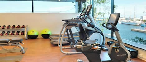 Une salle de sport équipée de tapis de course, d’haltères et de ballons d’exercice, avec vue sur un port de plaisance.