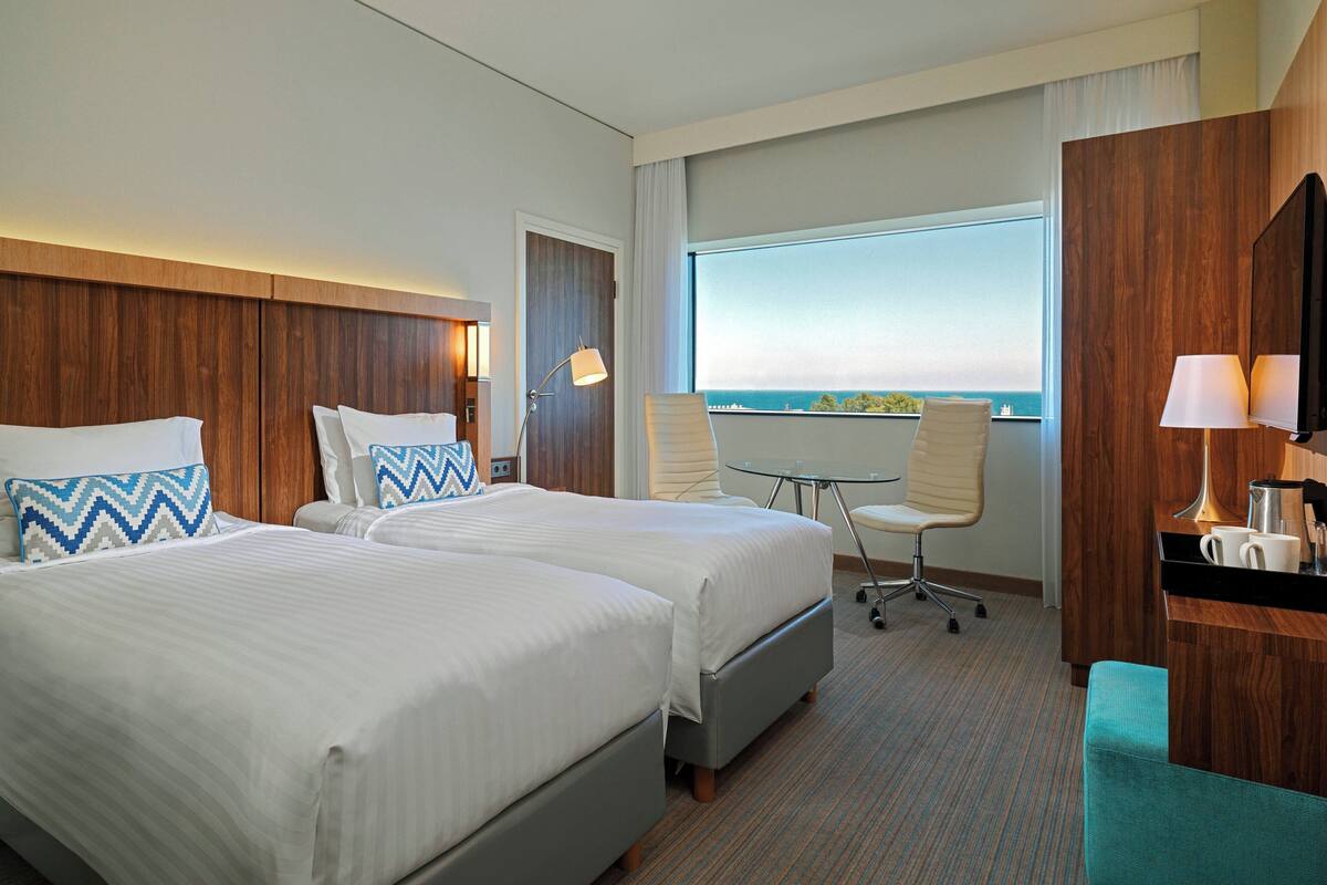 A hotel room with two beds, a desk, a chair, and a large window offering a view of the sea.