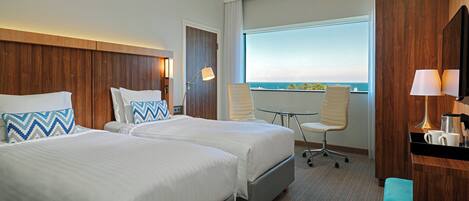 A hotel room with two beds, a desk, a chair, and a large window offering a view of the sea.