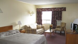 A hotel room with a bed, a sofa, a chair, a small table, a lamp, and a television. - Prospect Inn (Fredericton)
