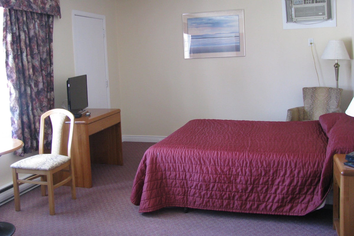 A hotel room with a bed, a desk with a computer, a chair, a lamp, and a painting on the wall.