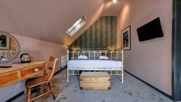 A bedroom with a sloped ceiling, a bed with white metal frame, a wooden desk with drawers, a chair, a small wooden chest, a radiator, and a mounted TV.
