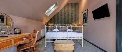 Een slaapkamer met een schuine kap, een bed met wit metalen frame, een houten bureau met lades, een stoel, een klein houten kistje, een radiator en een wandgemonteerde televisie.