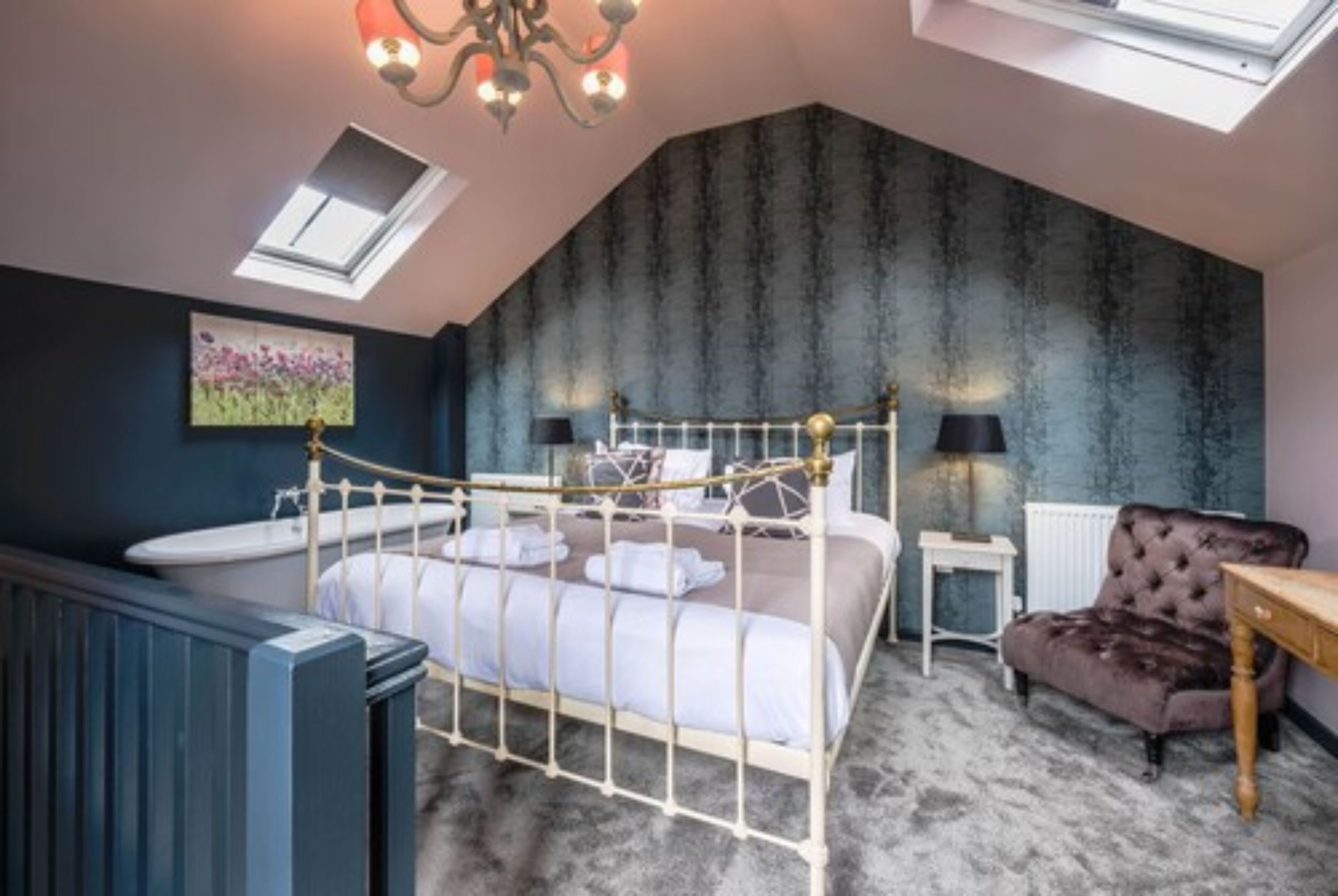 A bedroom with a sloped ceiling, a bed with a metal frame, a radiator, a small table, a chair, and a painting on the wall.