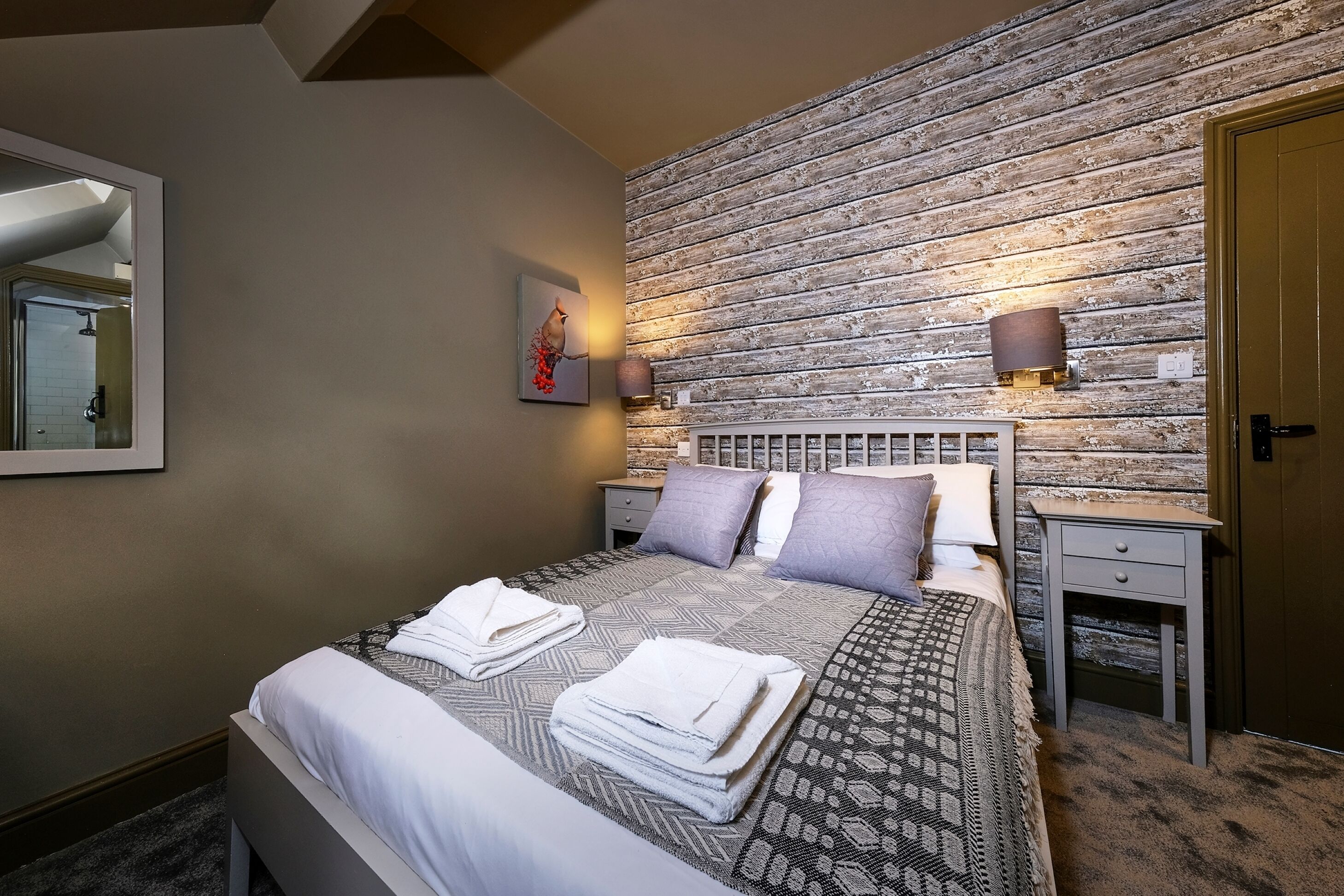 A neatly made bed with folded towels, a wooden textured wall, and a small bedside table with a lamp.