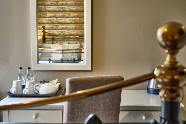 A hotel room with a dining table set with white cups, a teapot, and a tray. There is a framed picture on the wall and a lamp on the table.