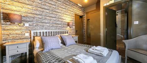 A hotel room with a rustic wooden wall, a bed with patterned bedding, a bedside table with a lamp, and a chair.