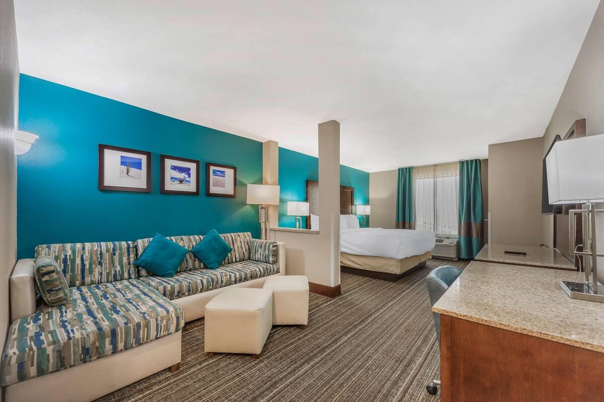 A hotel room with a teal accent wall, a sofa, a bed, a desk, and a small kitchenette.