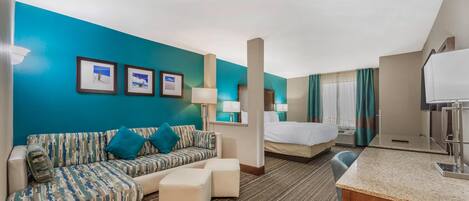 A hotel room with a teal accent wall, a sofa, a bed, a desk, and a small kitchenette.