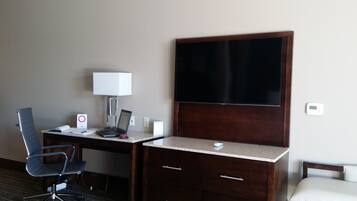A hotel room with a desk, chair, laptop, lamp, and a large flat-screen TV.